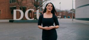 Video still featuring a woman talking and walking towards the camera with the DCU logo in the background