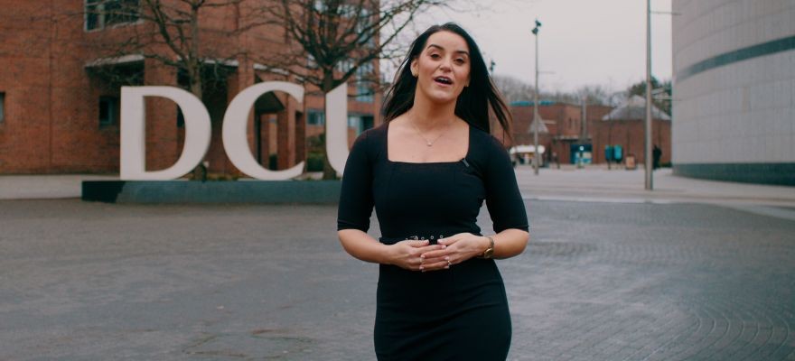 Video still featuring a woman talking and walking towards the camera with the DCU logo in the background